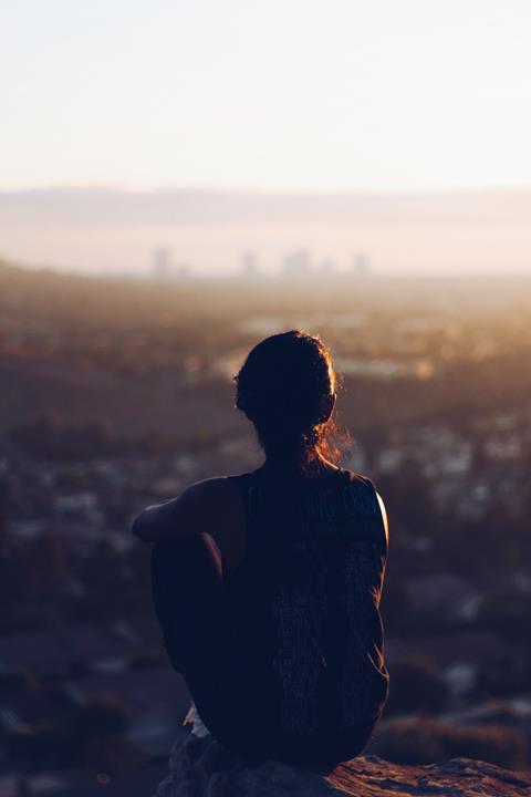 Observe - photo shows a silhouetted figure sat on a rock staring at a landscape; a cityscape in the backgroun out of focus.