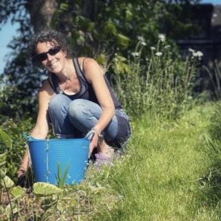 Katie, a white thin woman is wearing sunglasses and kneeling down in her garden with her hand on a blue bucket. she is smiling at the camera 