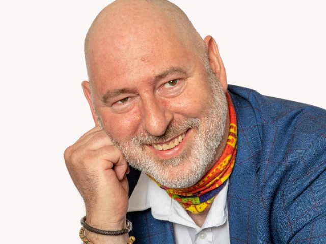 A picture of a man, in a smart suit jacket and neck scarf, with a short white beard smiling 