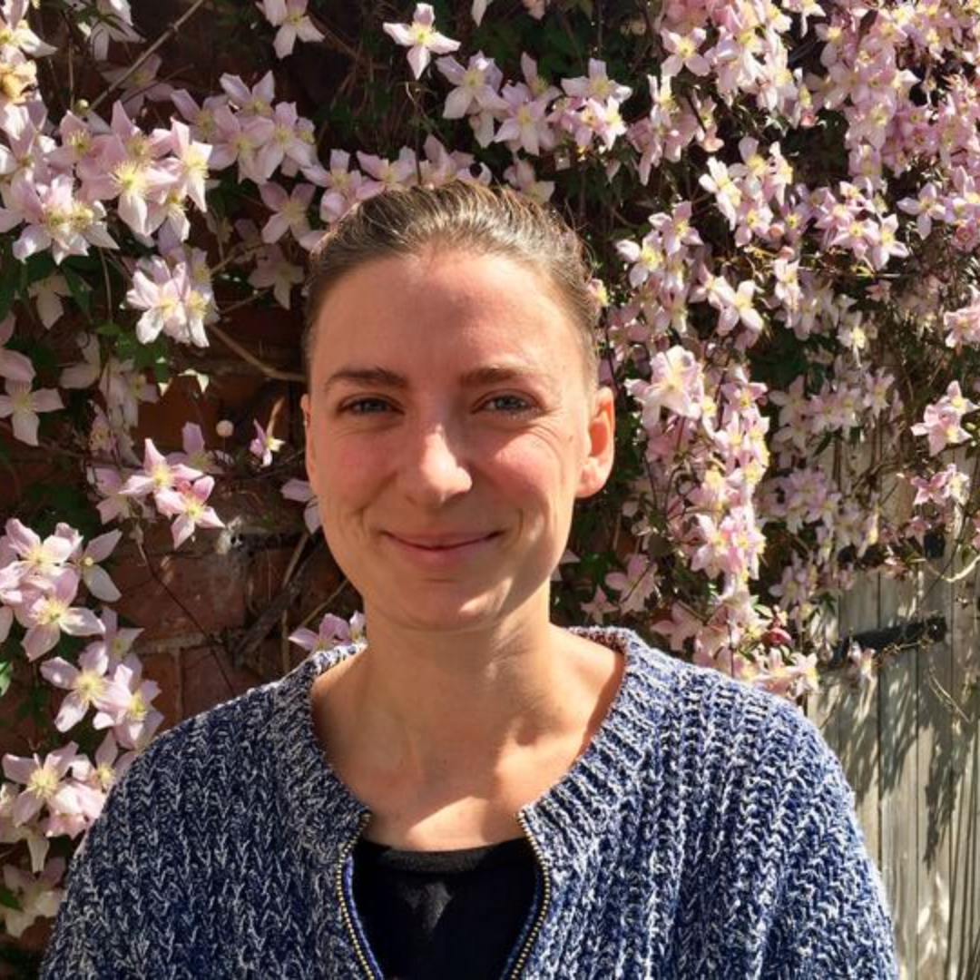 A white woman with brown hair pulled back off face into a ponytail. She is standing in front of a plant with pink flowers and is wearing a blue cardigan and black top. 