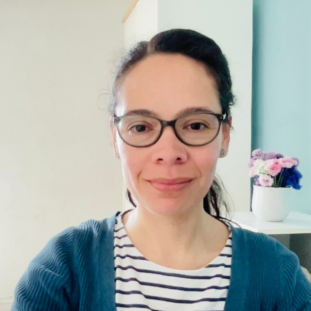 A photo of Maria - a woman with brown hair tied back and glasses. She is smiling. Wearing a blue and white stripy top and blue cardigan. 
