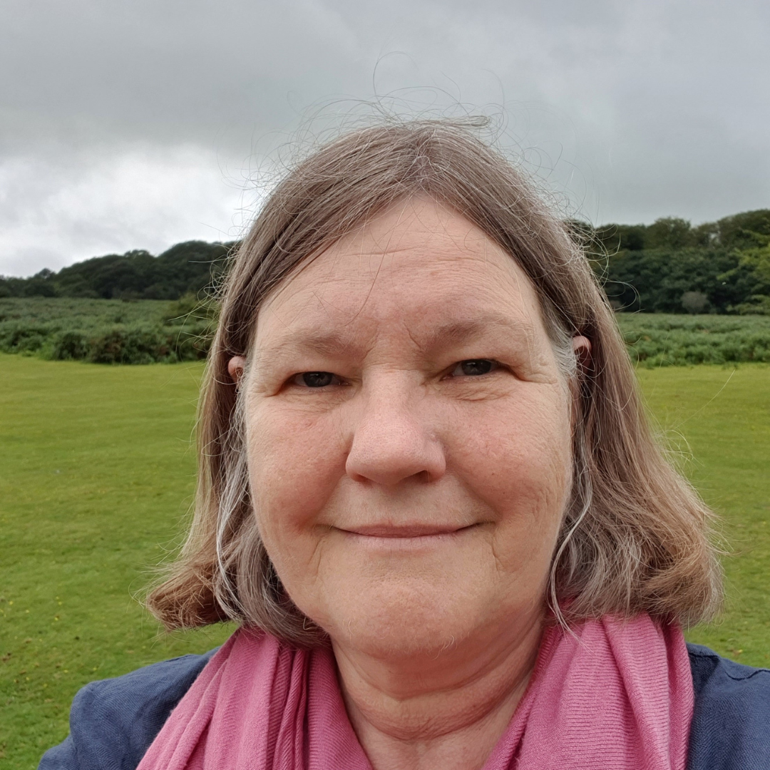 Image of a woman with a light brown bob, smiling. She is wearing a blue top and pink scarf and is outside in a field. 