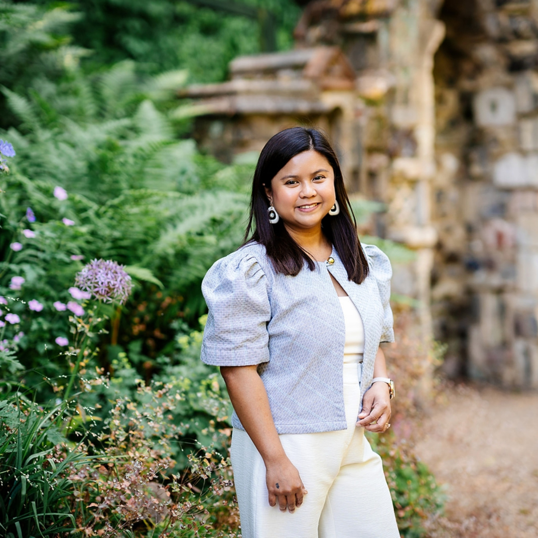Photo of a woman with long dark hair with the back drop of plants. She is wearing a light blue jacket and is smiling. 