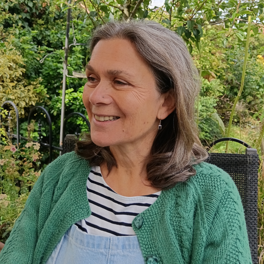 A picture of a smiling woman with shoulder length hair. She is sitting in a garden, wearing a blue and white stripy top and a green cardigan. 