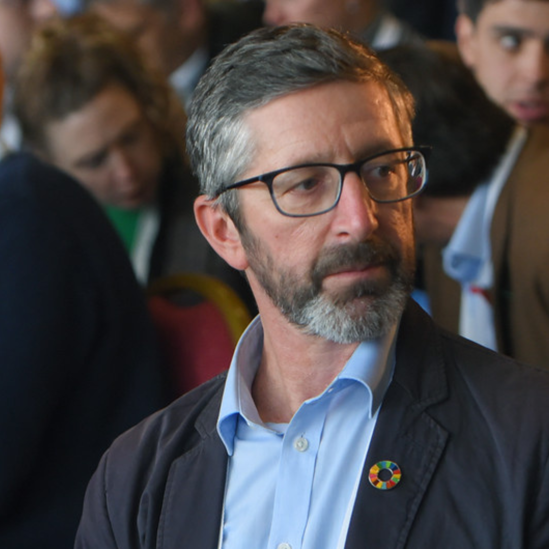 Image of a man with short grey hair, a beard and glasses. Wearing a blue suit. 