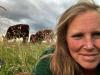 Photo of Hannah smiling at the camera with some of her Lincoln Red Cows behind her in long grass